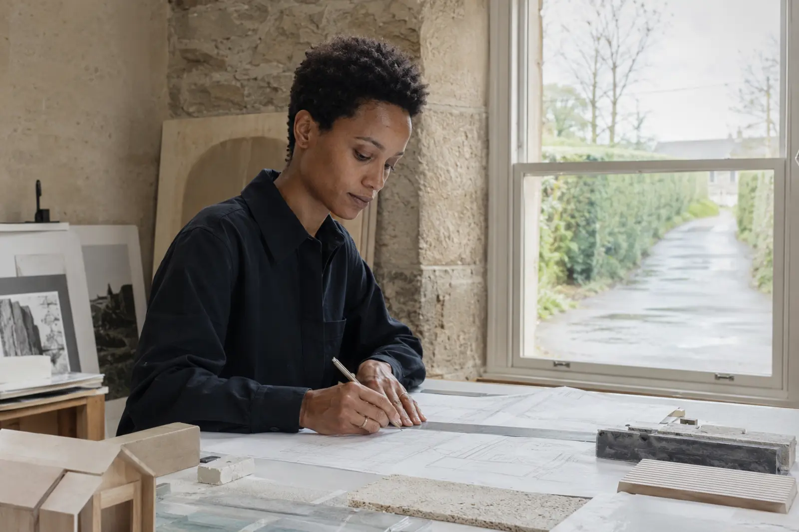 An architect sketching over drawings at a studio table with material samples and a timber model