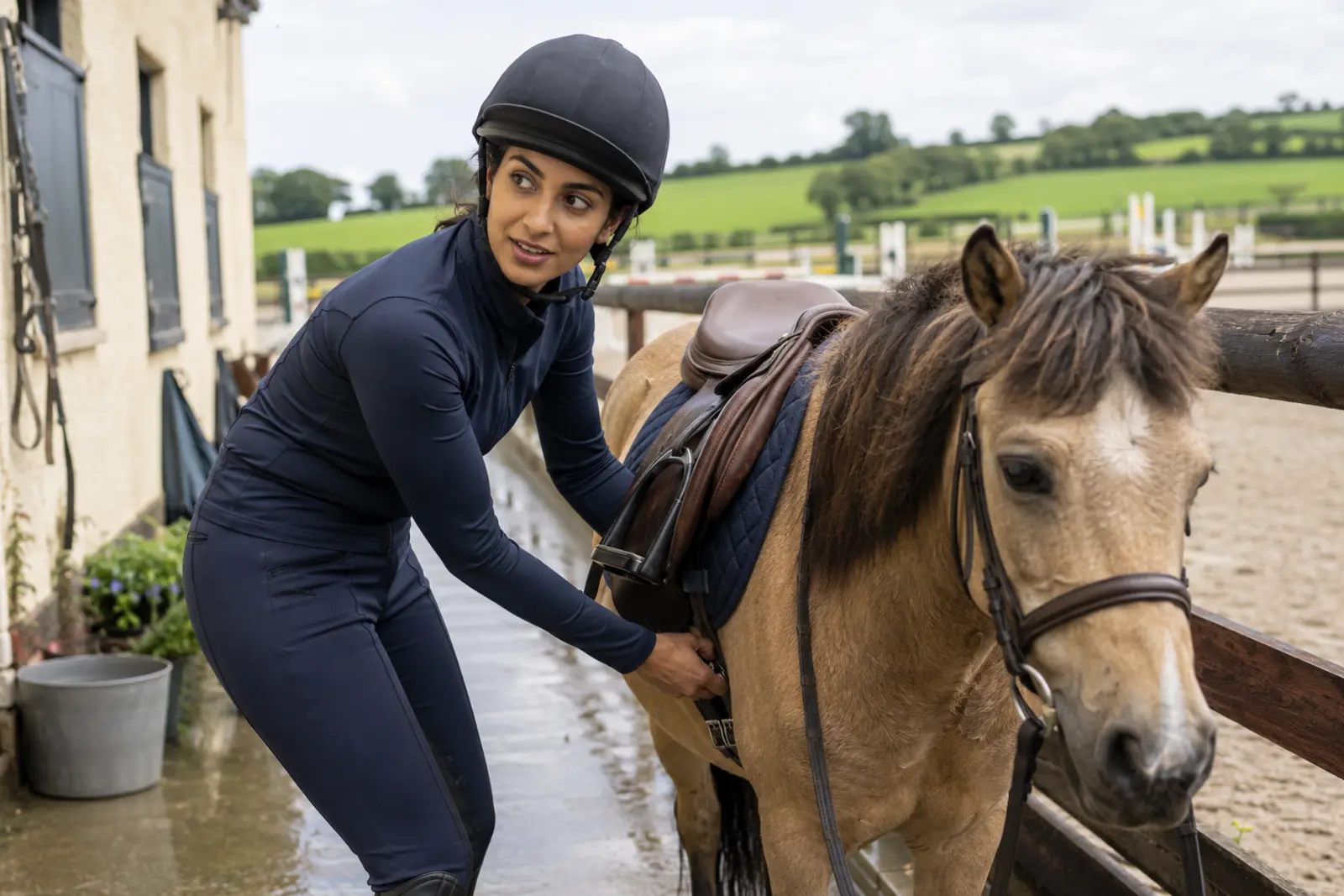 A rider in a helmet tightening the saddle on a dun pony by an outdoor arena