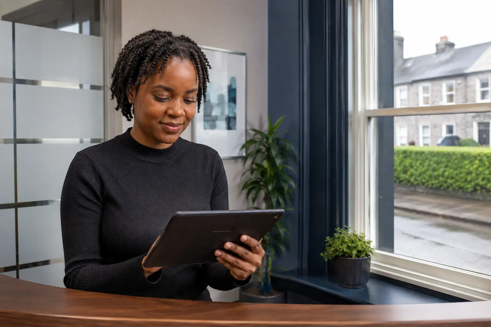 A clinic receptionist checking a tablet at the front desk by the window