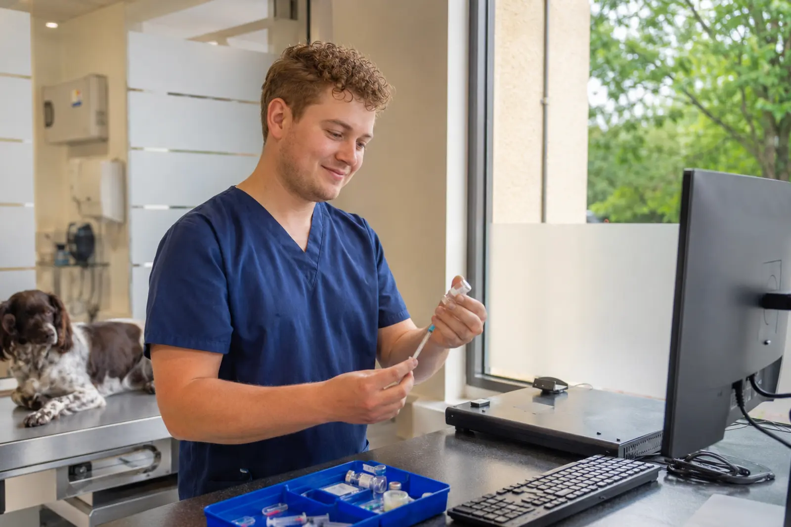 A vet preparing a vaccine at the counter with a spaniel resting on the table behind him