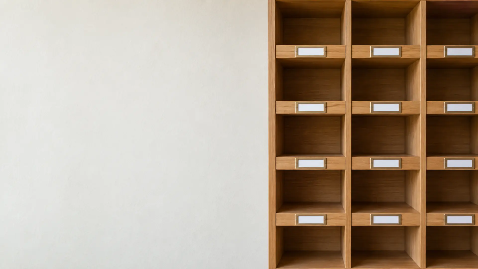 A wall of oak pigeonholes with blank brass label holders beside a plain plaster wall